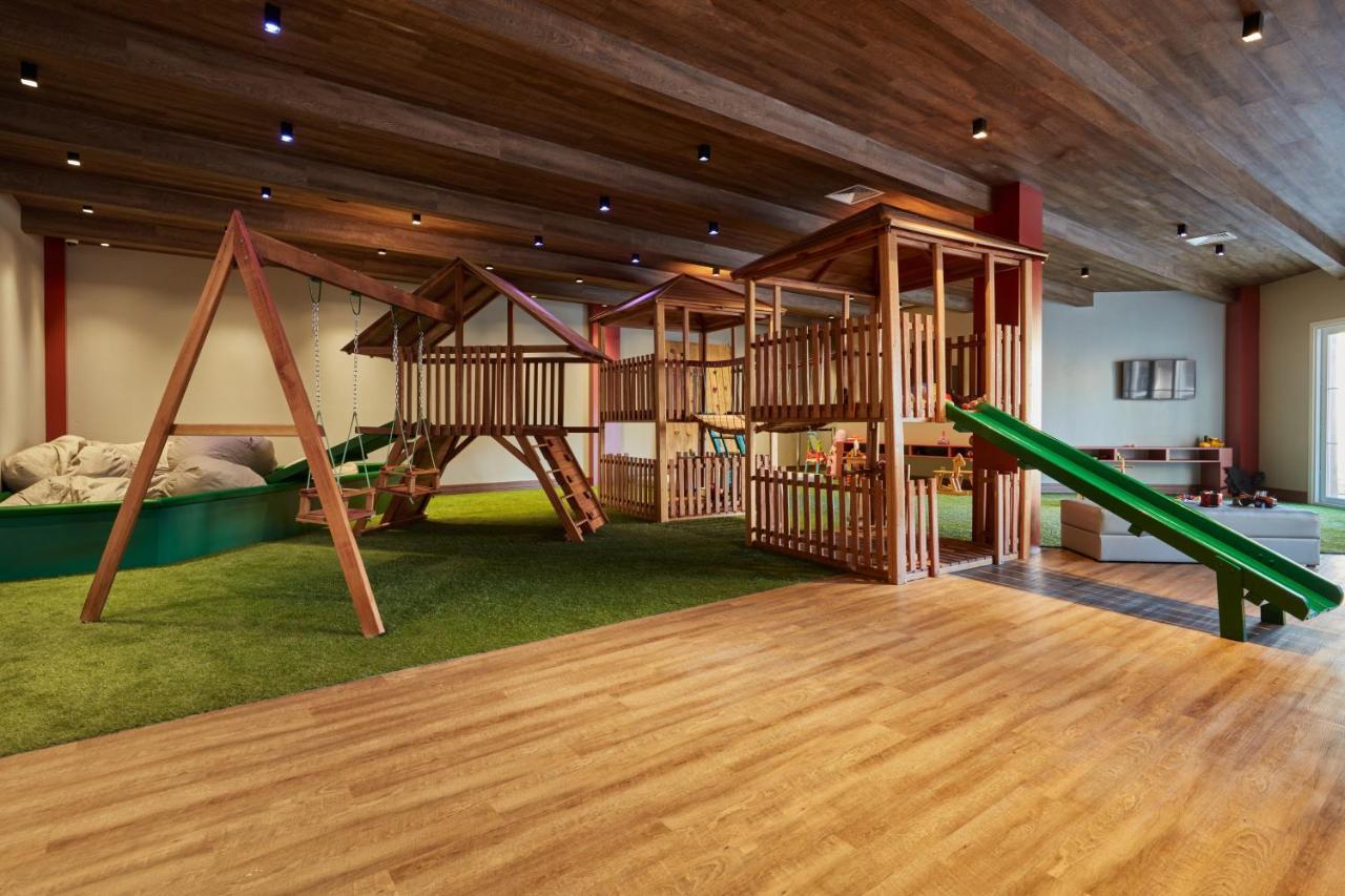 Buona Vitta Gramado Resort & Spa By Gramado Parks Buitenkant foto The photo shows an indoor play area designed for children. It features a wooden play structure that includes multiple levels, slides, and a swing set. The floor is covered with wooden planks, and there is a green carpet-like area that adds a soft tou