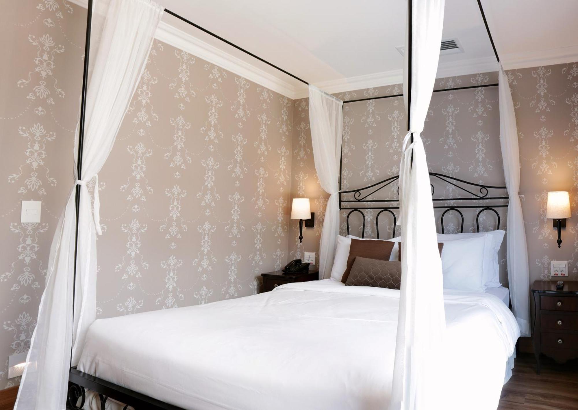 Buona Vitta Gramado Resort & Spa By Gramado Parks Buitenkant foto The photo shows a elegantly decorated bedroom featuring a four-poster bed with white drapes. The bed is made with white linens and has some brown accent pillows. The walls are adorned with a subtle floral pattern, and there are two bedside lamps prov