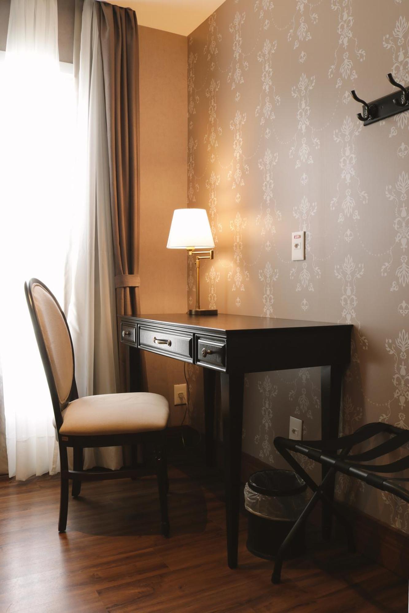 Buona Vitta Gramado Resort & Spa By Gramado Parks Buitenkant foto The photo shows a small desk set against a wall with a decorative pattern. The desk is black with a few drawers and has a small lamp on it. Next to the desk, there is a chair with a light-colored cushion. Soft light is coming through a window, castin