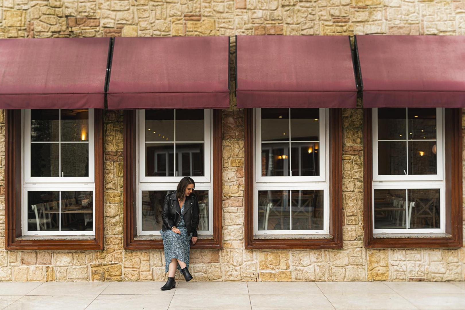 Buona Vitta Gramado Resort & Spa By Gramado Parks Buitenkant foto The photo shows a woman standing against a stone wall with three large windows in front of her, each framed with wooden borders. The windows have maroon awnings above them. The woman is dressed in a long skirt and leather jacket, leaning slightly aga